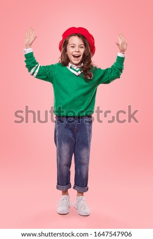 Full body excited child in colorful trendy outfit raising arms and looking at camera against pink background