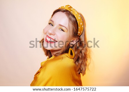 Beautiful young short-haired girl looking at the camera smiling. Girl with professional yellow make-up, bandage on her head, beautiful large earrings on a yellow background.