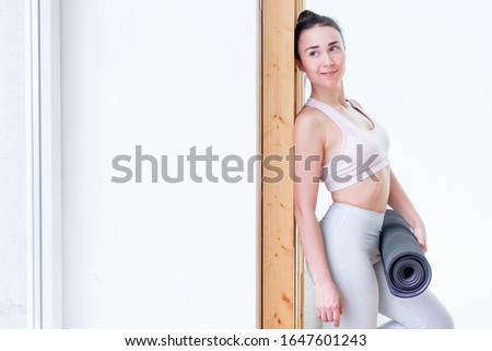 Beautiful young positive smiling caucasian woman pilates instructor with rug in her hands is standing in gym near window. Concept of regular classes. Advertising space