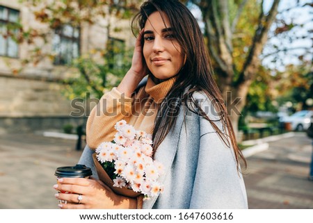 Brunette young woman drinking coffee on the go in the city street with a bouquet of flowers. Female in the street drinking hot beverage. Pretty brunette girl with cup of coffee in spring morning. 