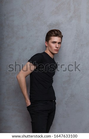 Young fashionable attractive man model with a stylish hairstyle in a trendy black t-shirt in vintage jeans with a nose piercing posing near a gray wall in the studio. Modern hipster guy indoors. Style