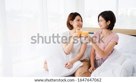 Two Asian women drink glass of orange juice in bedroom. Concept  for teenage or friends activity, lifestyle at home with copy space on the right side.