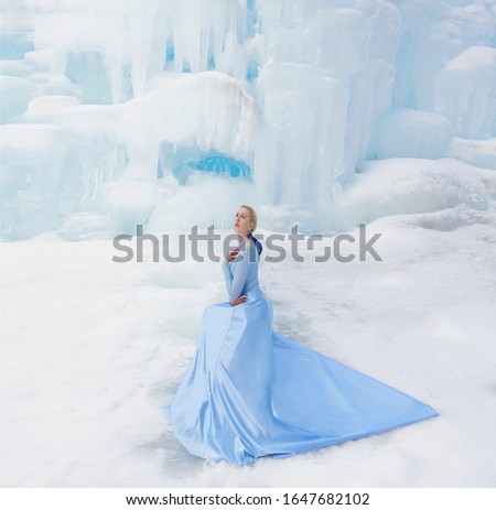 winter fairy tale on an ice waterfall a beautiful girl with long blonde hair and a blue dress with a long train.Snow Queen luxurious lush full dress waving silk fabric skirt long train.girl sit on ice