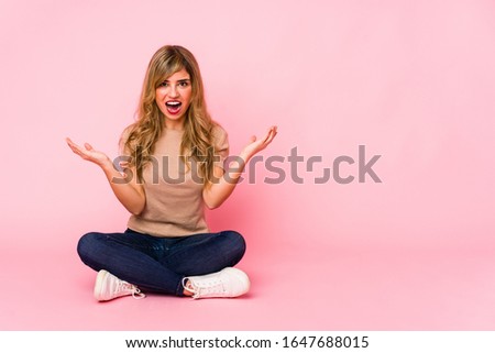 Young blonde caucasian woman sitting on a pink studio celebrating a victory or success, he is surprised and shocked.
