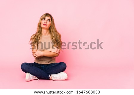 Young blonde caucasian woman sitting on a pink studio tired of a repetitive task.