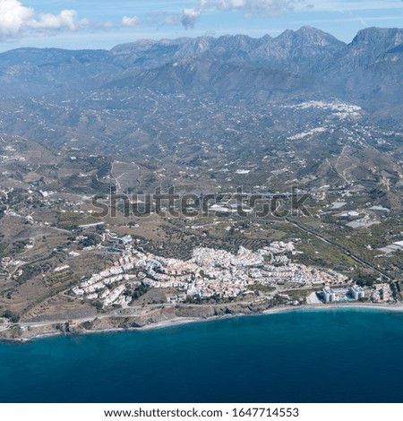 Aerial view of a small Spanisch beach town in Andalusia at the mediterranean coast. The sea is blue and clear. On the horizon a mountain ridge with blue sky and white clouds.