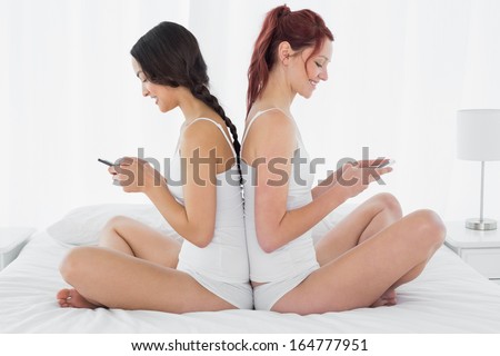 Side view of two smiling young female friends sitting back to back and text messaging on bed at home