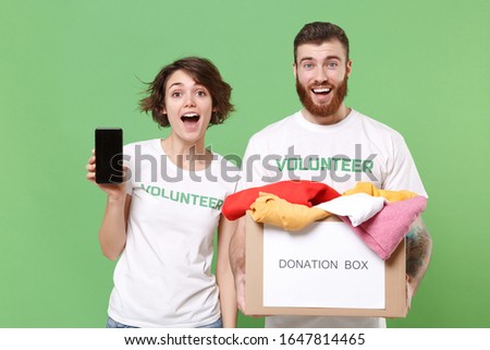 Excited friends couple in volunteer t-shirt isolated on green background. Voluntary free work assistance help charity grace teamwork. Hold donation box with clothes, mobile phone with empty screen