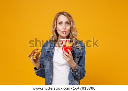 Pretty young woman in denim clothes isolated on orange background. Proper nutrition or American classic fast food concept. Mock up copy space. Hold burger french fries potatoes drink cola or soda