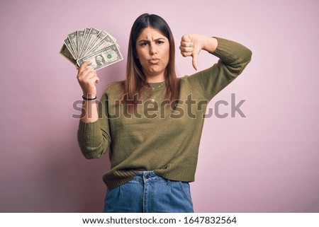 Young beautiful woman holding dollars standing over isolated pink background with angry face, negative sign showing dislike with thumbs down, rejection concept