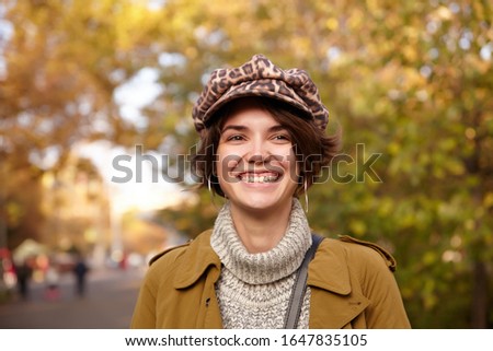 Closeup of beautiful attractive young brown haired lady with natural makeup laughing happily while walking over park on warm autumn day, dressed in stylish clothes