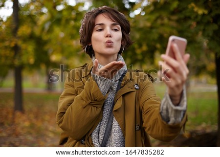 Charming young brown haired brunette lady with natural makeup folding her lips in air kiss and keeping her palm raised while making selfie with her mobile phone, posing outdoor on warm autumn day