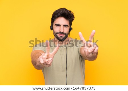 Telemarketer man working with a headset over isolated yellow background smiling and showing victory sign
