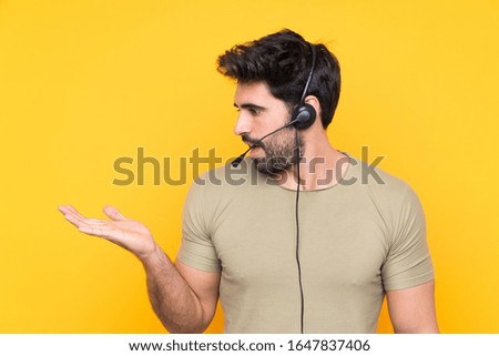 Telemarketer man working with a headset over isolated yellow background holding copyspace imaginary on the palm