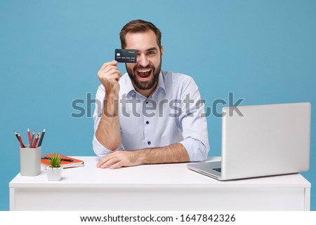 Funny young man in light shirt sit work at white desk with pc laptop isolated on pastel blue background. Achievement business career concept. Mock up copy space. Covering eye with credit bank card