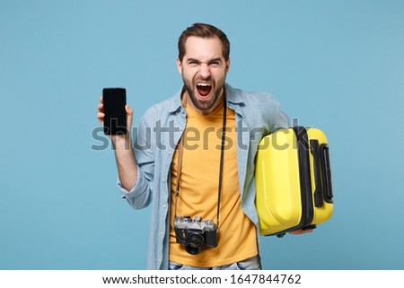 Screaming traveler tourist man in summer clothes with photo camera isolated on blue background. Passenger traveling abroad on weekends. Air flight journey Hold suitcase mobile phone with empty screen