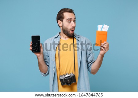 Amazed traveler tourist man in casual clothes with photo camera isolated on blue background. Passenger traveling on weekends. Air flight journey. Hold passport tickets mobile phone with empty screen
