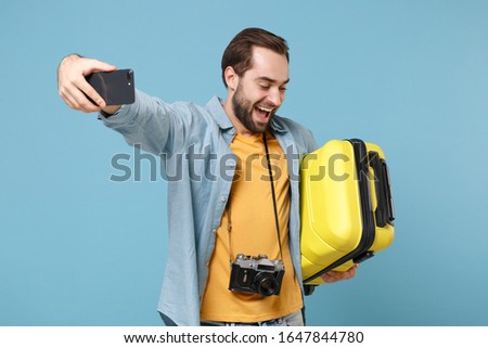 Joyful traveler tourist man in casual clothes with photo camera isolated on blue background. Passenger traveling abroad on weekends. Air flight journey Hold suitcase doing selfie shot on mobile phone