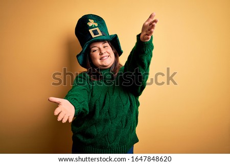Young beautiful plus size woman wearing green hat with clover celebrating saint patricks day looking at the camera smiling with open arms for hug. Cheerful expression embracing happiness.