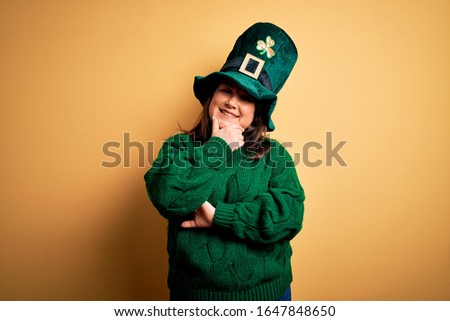 Young beautiful plus size woman wearing green hat with clover celebrating saint patricks day looking confident at the camera smiling with crossed arms and hand raised on chin. Thinking positive.