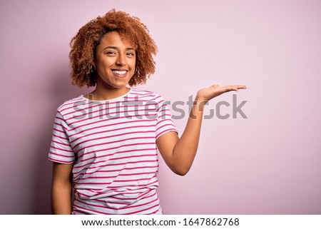 Young beautiful African American afro woman with curly hair wearing casual striped t-shirt smiling cheerful presenting and pointing with palm of hand looking at the camera.