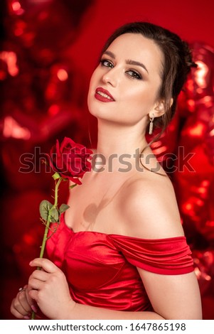 Beautiful young woman in red evening dress posing over red background with big heart shape balloons. 