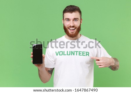 Excited man in volunteer t-shirt isolated on pastel green background. Voluntary free work assistance help charity grace teamwork concept. Pointing index finger on mobile phone with blank empty screen
