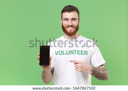 Smiling man in volunteer t-shirt isolated on pastel green background. Voluntary free work assistance help charity grace teamwork concept. Pointing index finger on mobile phone with blank empty screen