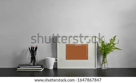 Photo of Pencil holder, Stack of books, Picture frame, Coffee cup and potted plant putting together on comfortable working table with living room as background. Orderly working space concept.