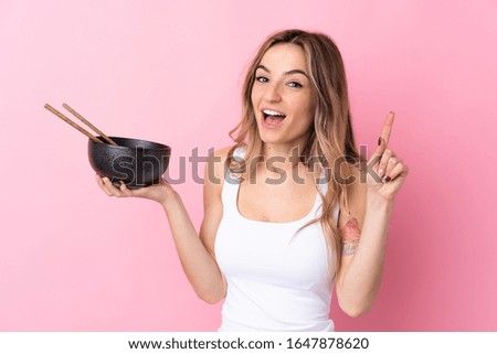 Young woman over isolated pink background pointing up a great idea while holding a bowl of noodles with chopsticks