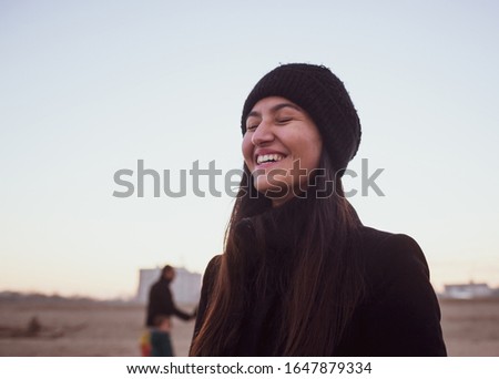 Young woman in winter clothing with wool hat smiling with closed eyes, emotion of authentic joy. Location on the beach with twilight light.