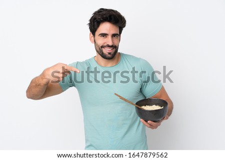 Young handsome man with beard over isolated white background and pointing it while holding a bowl of noodles with chopsticks