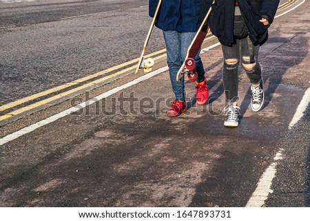 Two unrecognizable teenage girls carrying skateboards walking along a cycle path. One is wearing red shores the other has ripped black jeans.