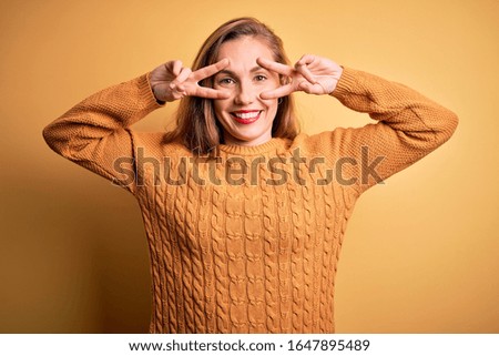 Young beautiful blonde woman wearing casual sweater standing over yellow background Doing peace symbol with fingers over face, smiling cheerful showing victory