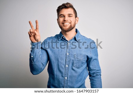 Young handsome blond man with beard and blue eyes wearing casual denim shirt smiling looking to the camera showing fingers doing victory sign. Number two.