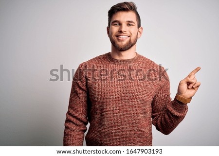 Young blond man with beard and blue eyes wearing casual sweater over white background with a big smile on face, pointing with hand finger to the side looking at the camera.