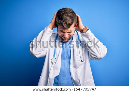 Young blond doctor man with beard and blue eyes wearing white coat and stethoscope suffering from headache desperate and stressed because pain and migraine. Hands on head.