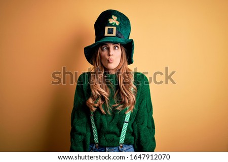 Beautiful brunette woman wearing green hat with clover celebrating saint patricks day making fish face with lips, crazy and comical gesture. Funny expression.