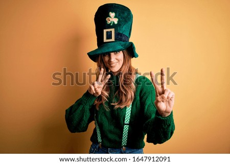 Beautiful brunette woman wearing green hat with clover celebrating saint patricks day smiling looking to the camera showing fingers doing victory sign. Number two.