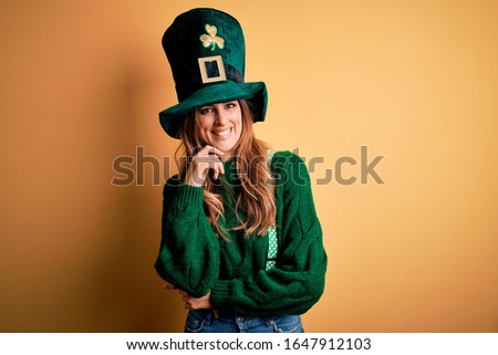 Beautiful brunette woman wearing green hat with clover celebrating saint patricks day looking confident at the camera with smile with crossed arms and hand raised on chin. Thinking positive.