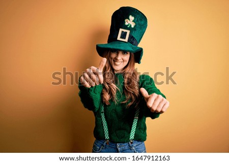 Beautiful brunette woman wearing green hat with clover celebrating saint patricks day approving doing positive gesture with hand, thumbs up smiling and happy for success. Winner gesture.