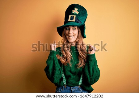 Beautiful brunette woman wearing green hat with clover celebrating saint patricks day celebrating surprised and amazed for success with arms raised and open eyes. Winner concept.