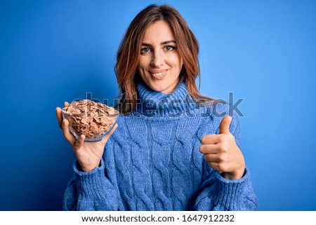 Young beautiful brunette woman holding bowl with healthy cornflakes cereals happy with big smile doing ok sign, thumb up with fingers, excellent sign