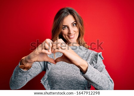 Young beautiful brunette woman wearing casual sweater standing over red background smiling in love showing heart symbol and shape with hands. Romantic concept.