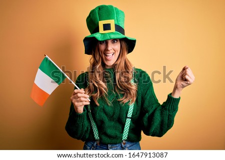 Beautiful patriotic woman wearing green hat holding irish flag celebrating saint patricks day screaming proud and celebrating victory and success very excited, cheering emotion
