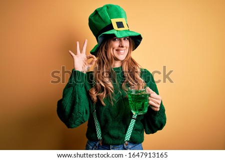 Beautiful woman wearing hat drinking jar of green beverage celebrating saint patricks day doing ok sign with fingers, excellent symbol