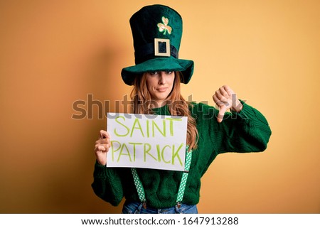 Beautiful brunette woman wearing green hat holding banner with saint patricks message with angry face, negative sign showing dislike with thumbs down, rejection concept