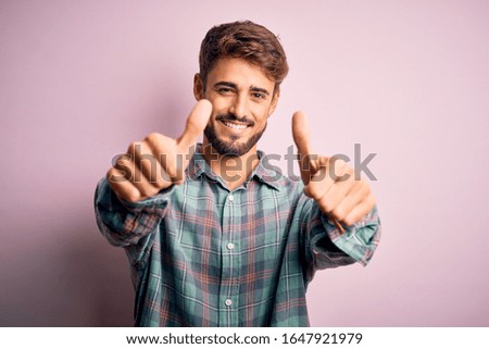 Young handsome man with beard wearing casual shirt standing over pink background approving doing positive gesture with hand, thumbs up smiling and happy for success. Winner gesture.