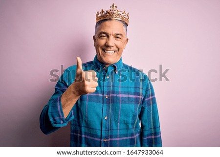 Middle age handsome grey-haired man wearing golden crown of king over pink background doing happy thumbs up gesture with hand. Approving expression looking at the camera showing success.