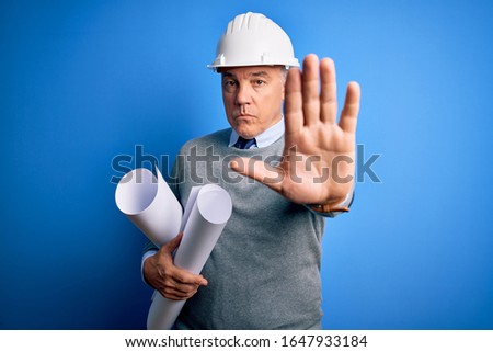 Middle age handsome grey-haired architect man wearing safety helmet holding blueprints doing stop sing with palm of the hand. Warning expression with negative and serious gesture on the face.
